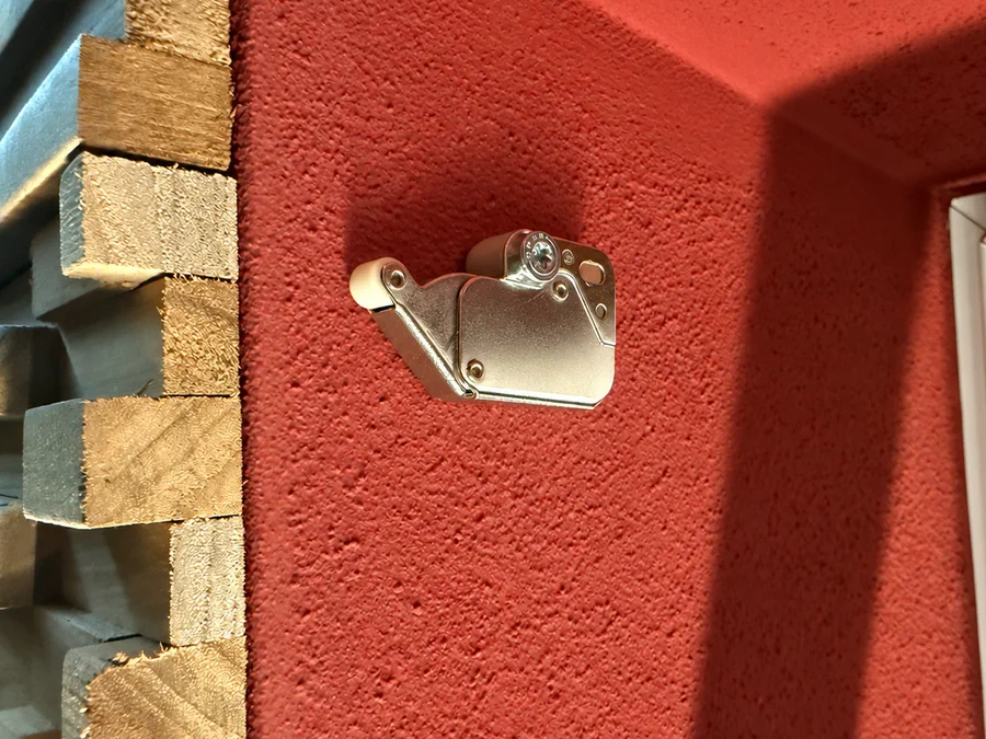 Metallischer Druckschnapper an der roten Fensterlaibung neben den Holzverblendern.
