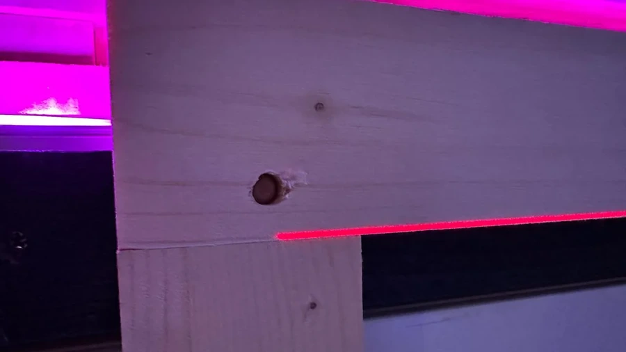 Nahaufnahme eines unbespannten Holzrahmens, den ein sichtbarer Holzdübel an der Unterkonstruktion hält; eine rote Laserlinie verläuft entlang der Rahmenkante