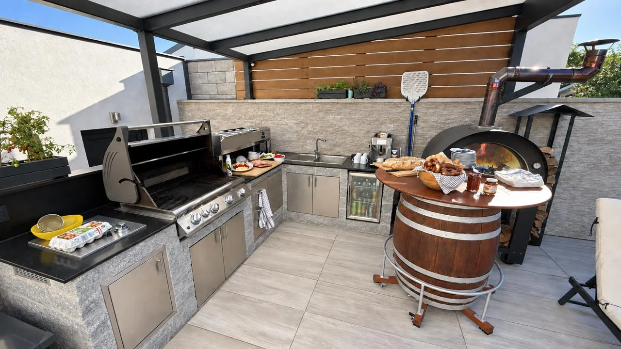 Outdoorküche während eines Brunchs mit geöffnetem Gasgrill, vorbereiteten Speisen, Spüle, Getränkekühlschrank und Holzbackofen