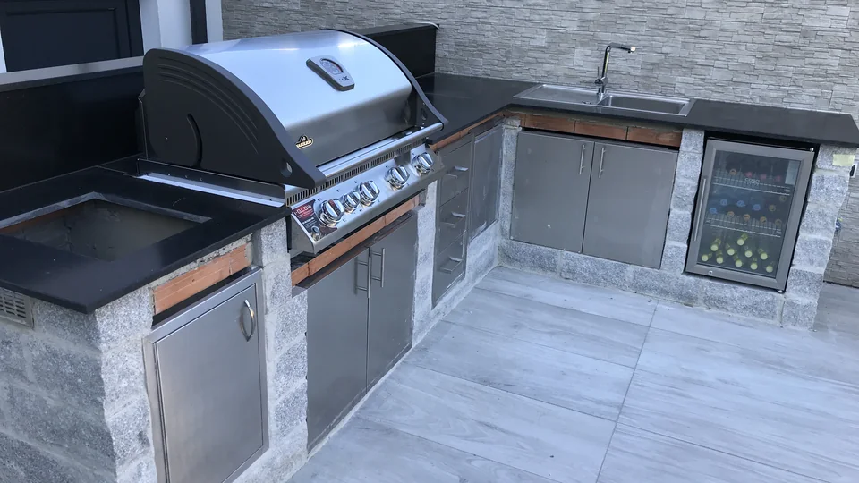 Outdoorküche mit montierter dunkler Arbeitsplatte, Grill, Spüle und Kühlschrank, aber noch sichtbaren orangefarbenen Auflagern