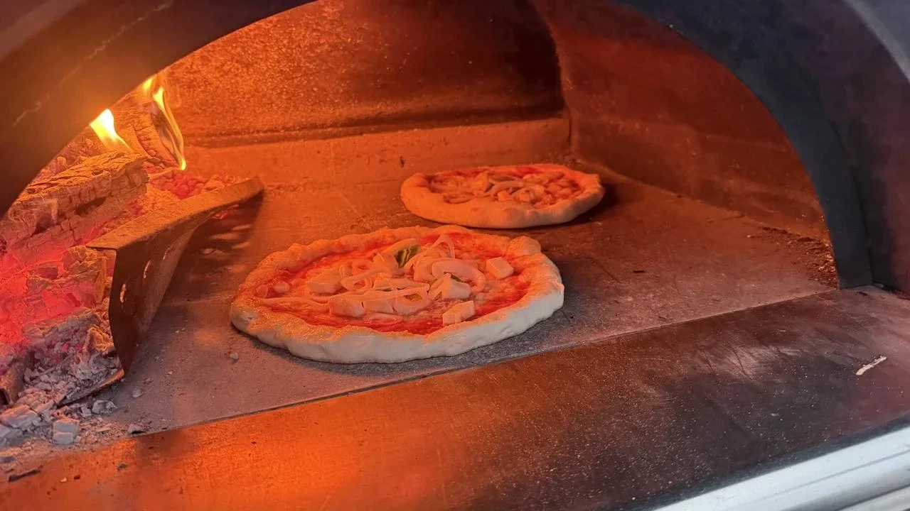 Zwei Pizzen im Holzofen, links daneben glühendes Holz und sichtbare Flammen.