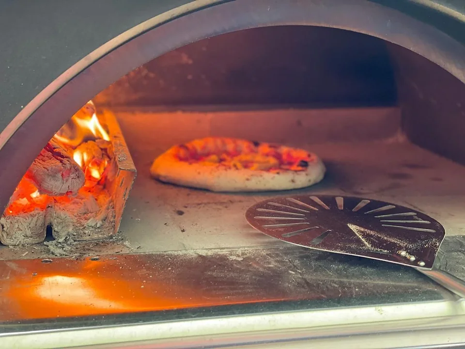 Pizza beim Backen auf dem Stein im Holzofen, links brennendes Holz und vorne ein Pizzawender
