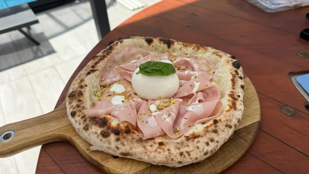 Pizza mit Mortadella, gehackten Pistazien, weißem Käse und Basilikum auf einem Holzbrett.