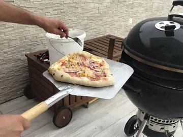 Pizza mit gebräuntem Rand auf einer Pizzaschaufel neben einem schwarzen Kugelgrill.