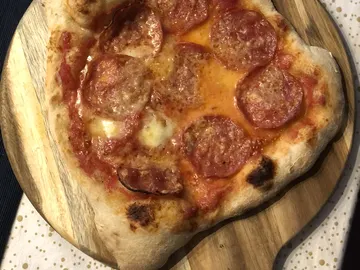 Unregelmäßig geformte Pizza mit sichtbaren Salamischeiben auf einem runden Holzbrett.