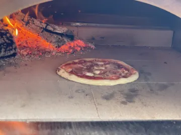 Pizza auf der Steinfläche des Holzofens, links daneben Glut und brennendes Holz.