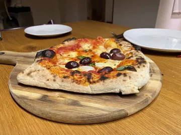 Unregelmäßig geformte Pizza mit schwarzen Oliven auf einem Holzbrett.