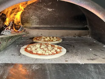 Zwei Pizzen hintereinander auf dem Steinboden des Holzofens, links daneben brennendes Holz und Flammen.