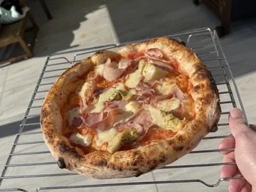 Eine Hand hält ein Metallgitter mit einer Pizza mit Schinken und Artischocken.