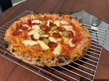 Pizza mit Salami, grünen Oliven und Käse auf einem Metallgitter.