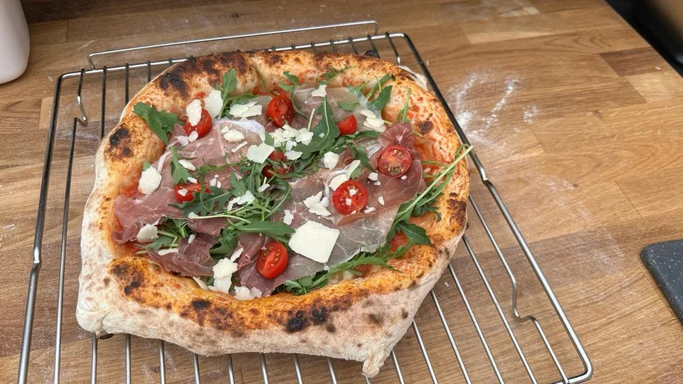 Frisch gebackene Pizza mit Prosciutto, Rucola, Kirschtomaten und gehobeltem Hartkäse auf einem Abkühlgitter.