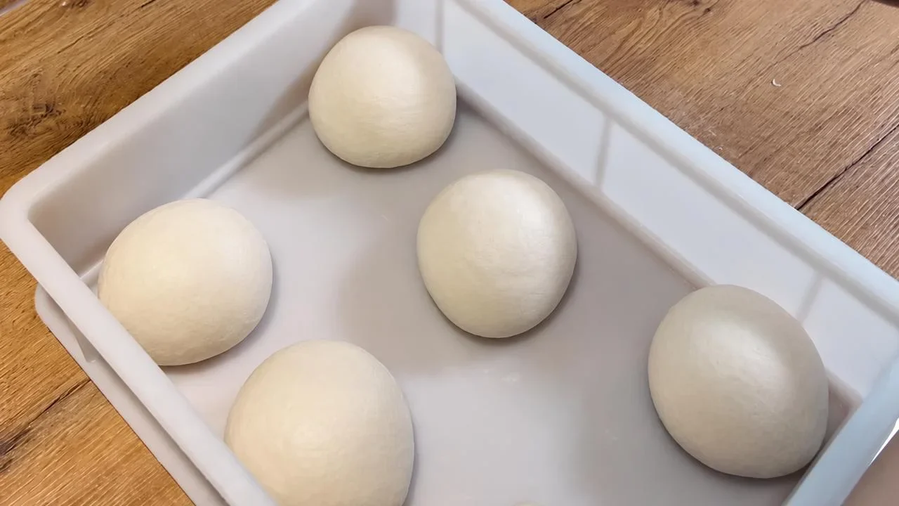 Mehrere glatte Pizzateigballen mit Abstand in einer weißen Teigbox auf einem Holztisch.