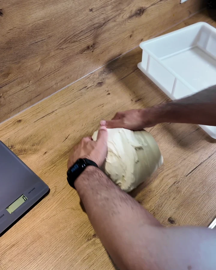 Mario schleift die gesamte Pizzateigmasse mit beiden Händen auf einer Holzarbeitsplatte rund.