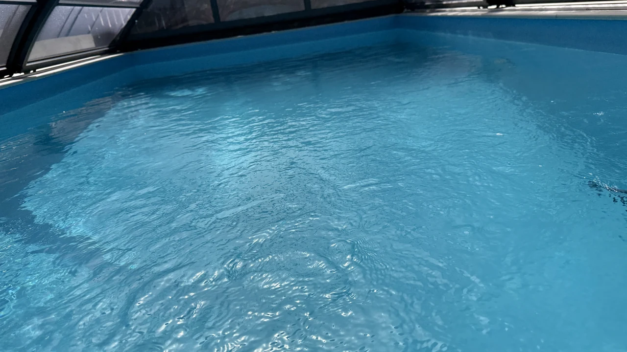 Blaues Poolwasser mit Sonnenreflexen unter der teilweise geöffneten dunklen Poolüberdachung.