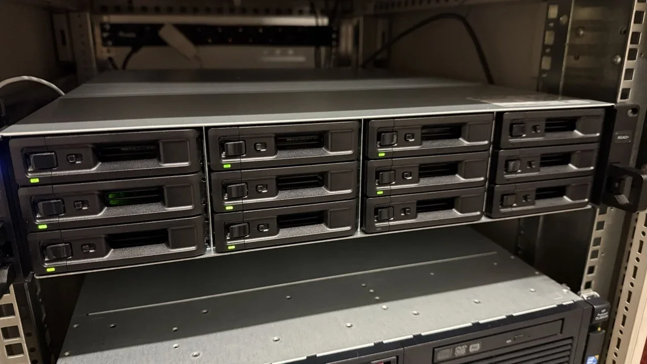 Leicht schräge Frontansicht der Synology RS2423+ mit zwölf Laufwerkseinschüben und grün leuchtenden Status-LEDs in einem Serverschrank.
